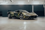 A sleek, modern sports car with a glossy olive green exterior is parked in a spacious indoor garage. The car's design features sharp, aerodynamic lines and distinctive curves. A badge on the side suggests a luxury brand, and the car sits on stylish alloy wheels. The garage has a polished floor and utilitarian lighting.