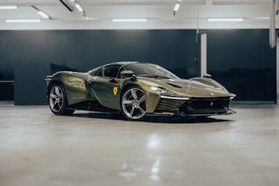 A sleek, modern sports car with a glossy olive green exterior is parked in a spacious indoor garage. The car's design features sharp, aerodynamic lines and distinctive curves. A badge on the side suggests a luxury brand, and the car sits on stylish alloy wheels. The garage has a polished floor and utilitarian lighting.