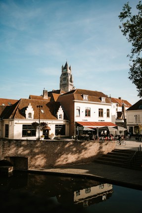 A picturesque European street scene with a tall, historic tower in the background. The foreground features charming buildings with terracotta roofs, a caf&eacute; with outdoor seating under a red and white awning, and bicycles parked nearby. The sky is clear, and the setting sun casts warm shadows.