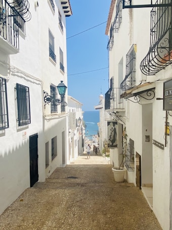 A narrow cobblestone street is flanked by white-washed buildings with black wrought iron balconies. The bright blue sky and distant sea add a serene atmosphere. A couple of people are walking down towards the water, enhancing the sense of leisure and exploration. Signage hangs from the sides of the buildings, suggesting this is a quaint, possibly historic area.