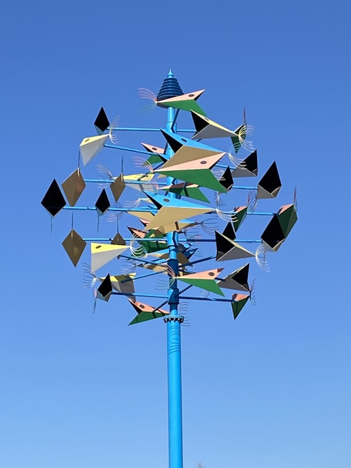 A colorful kinetic sculpture with multiple geometric shapes mounted on a tall blue pole. The shapes have a mix of triangular, diamond, and rectangular forms in various colors with tails, arranged to rotate and move in the wind.