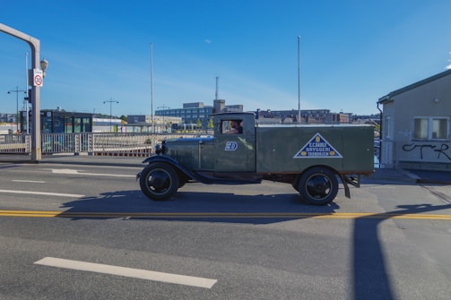 A vintage green delivery truck is driving on a road in an urban setting. The truck has a logo on the side with text, and a driver is visible inside the cabin. The background includes modern buildings, a bridge, and clear blue skies.