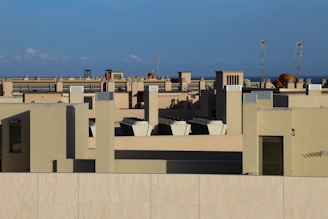A rooftop landscape featuring various tan building structures with multiple ventilation units and other technical equipment. In the background, blue sky and distant antennas can be observed.
