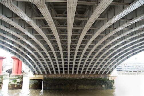 The image depicts the underside of a metal bridge with a series of evenly spaced, arched steel beams. Two large red cylindrical columns support the bridge, and a river flows beneath it. The surrounding area appears industrial.