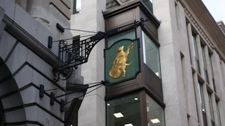 A decorative sign attached to a building features a gold cat playing a cello. The sign is mounted on a wrought iron frame and placed above a contemporary building façade with large windows. The architecture combines classic and modern styles, using stone and metal elements.