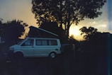 A camper van with its roof extended is parked in a natural setting, surrounded by trees. The sun is either setting or rising, casting a warm glow on the horizon, creating a serene atmosphere. The overall scene appears tranquil with the silhouettes of trees and the van prominently visible.