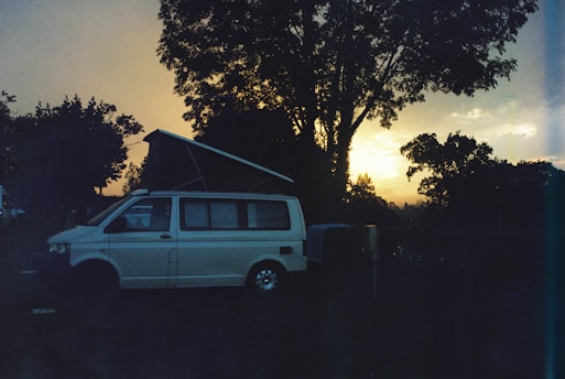 A camper van with its roof extended is parked in a natural setting, surrounded by trees. The sun is either setting or rising, casting a warm glow on the horizon, creating a serene atmosphere. The overall scene appears tranquil with the silhouettes of trees and the van prominently visible.