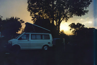 A cozy camper van parked by a river surrounded by tall trees under a warm sunset sky.