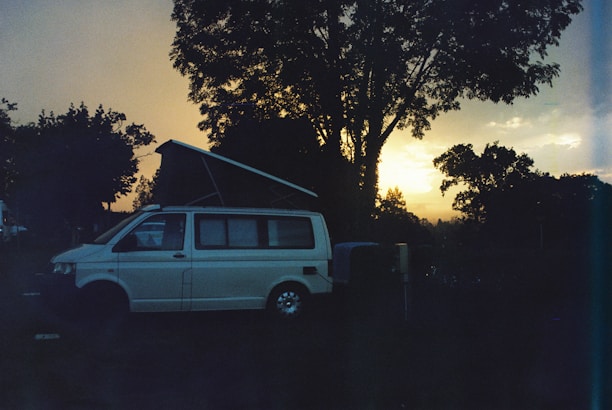 A camper van with its roof extended is parked in a natural setting, surrounded by trees. The sun is either setting or rising, casting a warm glow on the horizon, creating a serene atmosphere. The overall scene appears tranquil with the silhouettes of trees and the van prominently visible.