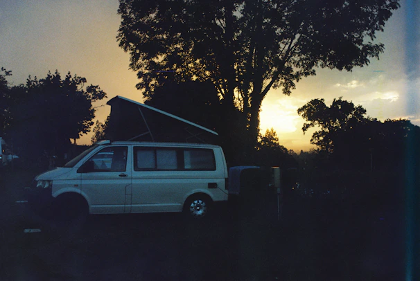 A sleek camper van parked by a serene forest road at sunset, with warm orange and green hues.