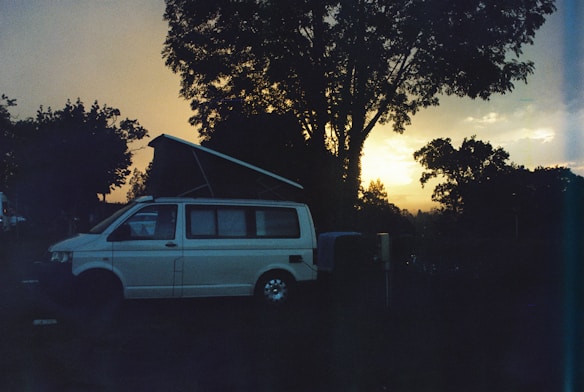 A camper van with its roof extended is parked in a natural setting, surrounded by trees. The sun is either setting or rising, casting a warm glow on the horizon, creating a serene atmosphere. The overall scene appears tranquil with the silhouettes of trees and the van prominently visible.
