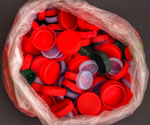 A plastic bag is filled with numerous colorful bottle caps, primarily red, with a few green and purple caps mixed in. The caps are tightly packed within the translucent bag.