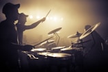 A band is performing energetically on stage with a drummer in the foreground. The stage is lit with bright lights creating a dramatic silhouette effect. The audience is faintly visible in the background, contributing to the lively atmosphere.
