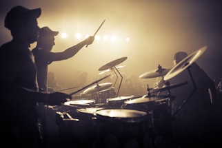 A band is performing energetically on stage with a drummer in the foreground. The stage is lit with bright lights creating a dramatic silhouette effect. The audience is faintly visible in the background, contributing to the lively atmosphere.