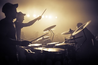 A band is performing energetically on stage with a drummer in the foreground. The stage is lit with bright lights creating a dramatic silhouette effect. The audience is faintly visible in the background, contributing to the lively atmosphere.