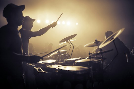 A band is performing energetically on stage with a drummer in the foreground. The stage is lit with bright lights creating a dramatic silhouette effect. The audience is faintly visible in the background, contributing to the lively atmosphere.