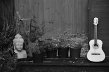 A serene outdoor scene featuring a stone sculpture of a Buddha head surrounded by various potted plants. Beside the plants, a classical guitar rests against a wooden panel or fence. The entire setting has a calm and natural atmosphere, enhanced by the monochrome color scheme.