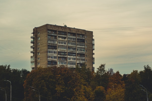 A tall, rectangular apartment building stands amid a dense area of trees with multiple floors and balconies. The sky appears overcast, giving the surroundings a muted and subdued tone.