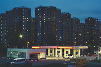 A gas station is illuminated at night, with a few vehicles parked nearby. Tall apartment buildings form the backdrop, their windows glowing warmly against the dusk sky.