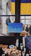 A scene inside a building with a Ukrainian flag hanging on the wall. A painting of a blue sky with some clouds is propped up against a window. On a table, there are sandwiches wrapped in plastic, pastries, paper cups, and other items. A group of people is visible in the background through the window.