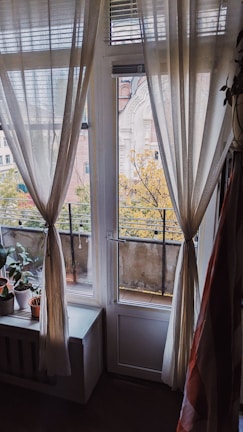 Curtains gracefully hang around a glass door and window leading to a balcony, with a view of autumn trees and an old building. Potted plants are placed on a windowsill inside a warmly lit room, adding a homely touch.