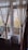 Curtains gracefully hang around a glass door and window leading to a balcony, with a view of autumn trees and an old building. Potted plants are placed on a windowsill inside a warmly lit room, adding a homely touch.