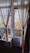 Curtains gracefully hang around a glass door and window leading to a balcony, with a view of autumn trees and an old building. Potted plants are placed on a windowsill inside a warmly lit room, adding a homely touch.