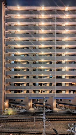 A tall residential building with multiple stories features rows of balconies lit by warm lights. The structure is made of brick and concrete. At the base, a row of parked bicycles is visible near the railway tracks, which run parallel to the building.