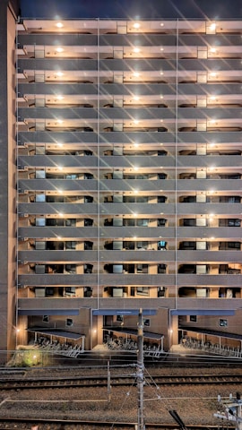 A tall residential building with multiple stories features rows of balconies lit by warm lights. The structure is made of brick and concrete. At the base, a row of parked bicycles is visible near the railway tracks, which run parallel to the building.