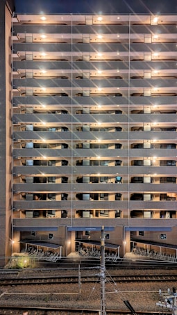 A tall residential building with multiple stories features rows of balconies lit by warm lights. The structure is made of brick and concrete. At the base, a row of parked bicycles is visible near the railway tracks, which run parallel to the building.