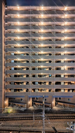 A tall residential building with multiple stories features rows of balconies lit by warm lights. The structure is made of brick and concrete. At the base, a row of parked bicycles is visible near the railway tracks, which run parallel to the building.