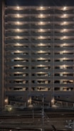 A multi-story residential building at night with exterior hallway lights illuminating each floor. The lights create a symmetrical pattern, highlighting the structure's repetitive architectural features. Below, there is a bike storage area, and train tracks are visible in the foreground running parallel to the building.