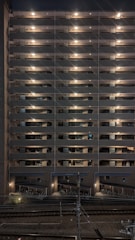 A multi-story residential building at night with exterior hallway lights illuminating each floor. The lights create a symmetrical pattern, highlighting the structure's repetitive architectural features. Below, there is a bike storage area, and train tracks are visible in the foreground running parallel to the building.