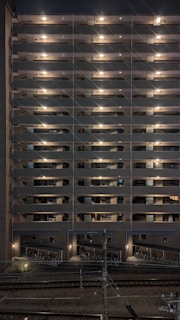 A multi-story residential building at night with exterior hallway lights illuminating each floor. The lights create a symmetrical pattern, highlighting the structure's repetitive architectural features. Below, there is a bike storage area, and train tracks are visible in the foreground running parallel to the building.