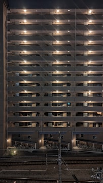 A multi-story residential building at night with exterior hallway lights illuminating each floor. The lights create a symmetrical pattern, highlighting the structure's repetitive architectural features. Below, there is a bike storage area, and train tracks are visible in the foreground running parallel to the building.