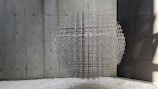 A spherical sculpture composed of interwoven metal rods is suspended in a minimalist environment with concrete walls. The sculpture casts complex shadows on the surrounding surfaces. The floor is covered with white gravel, adding texture to the setting.