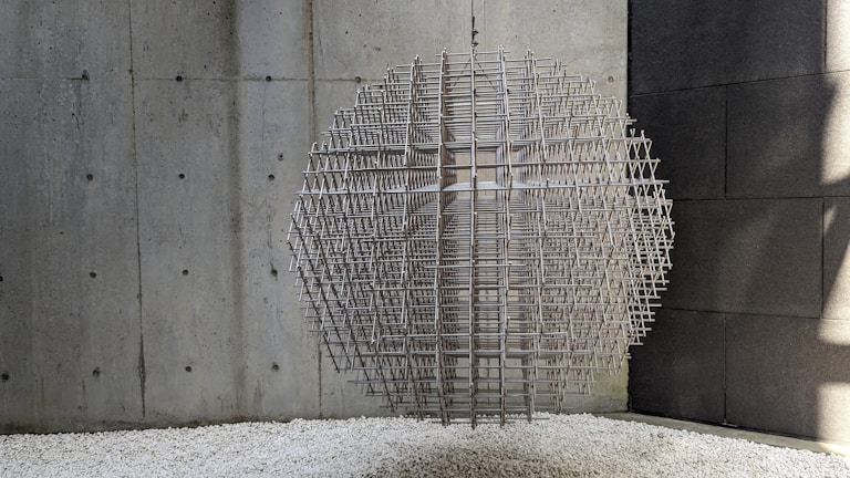 A spherical sculpture composed of interwoven metal rods is suspended in a minimalist environment with concrete walls. The sculpture casts complex shadows on the surrounding surfaces. The floor is covered with white gravel, adding texture to the setting.