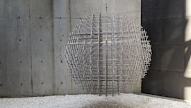 A spherical sculpture composed of interwoven metal rods is suspended in a minimalist environment with concrete walls. The sculpture casts complex shadows on the surrounding surfaces. The floor is covered with white gravel, adding texture to the setting.