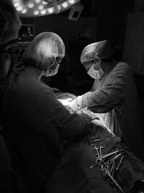 Medical professionals in scrubs and masks are performing surgery under bright surgical lights. A variety of surgical instruments are organized on a covered tray next to the operating table.