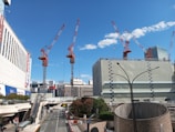 A cityscape featuring several large cranes, indicating ongoing construction. Tall buildings surround the area, with a clear blue sky overhead accented by a few clouds. The street below includes a bus, trees, and pedestrians. A modern, urban setting with a focus on development.