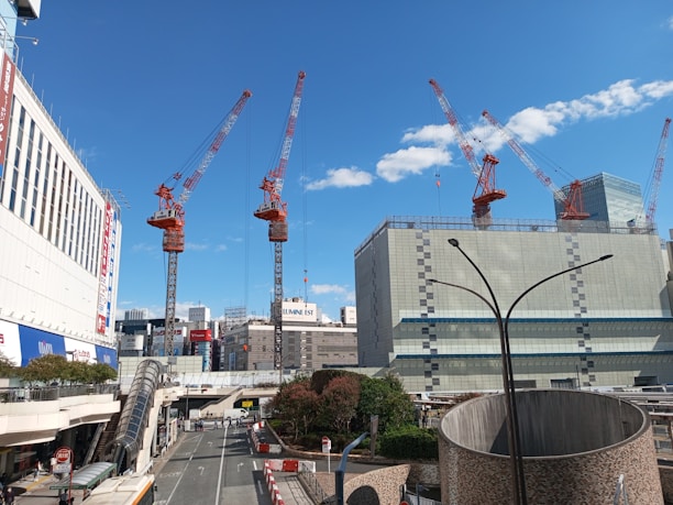 A cityscape featuring several large cranes, indicating ongoing construction. Tall buildings surround the area, with a clear blue sky overhead accented by a few clouds. The street below includes a bus, trees, and pedestrians. A modern, urban setting with a focus on development.