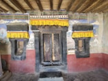 A rustic wooden doorway flanked by two windows, all surrounded by stone walls. The top features colorful yellow fabric canopies with decorative designs. The wall is adorned with faded mural art, blending hues of blue, red, and green.
