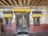 A rustic wooden doorway flanked by two windows, all surrounded by stone walls. The top features colorful yellow fabric canopies with decorative designs. The wall is adorned with faded mural art, blending hues of blue, red, and green.
