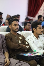 A group of people seated in an auditorium setting. The focus is on two men in the foreground, one wearing a brown shirt with leaf patterns, holding a video game controller with colorful buttons. Others are sitting in the background.