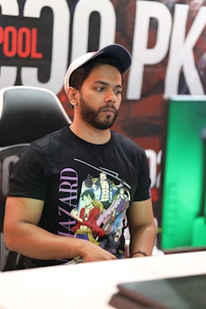A man with a beard sits in a chair wearing a black t-shirt featuring colorful anime characters. He has a focused expression and is wearing a white cap backwards. In the background, there are blurred, colorful elements and text in red and white.