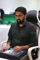 A person is seated in a chair, focused intently on something, with hands positioned on a gaming console or arcade controller. The setup appears to be indoors, with visible screens and lighting in the background, suggesting an electronic gaming environment.