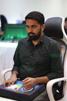 A person is seated in a chair, focused intently on something, with hands positioned on a gaming console or arcade controller. The setup appears to be indoors, with visible screens and lighting in the background, suggesting an electronic gaming environment.