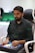 A person is seated in a chair, focused intently on something, with hands positioned on a gaming console or arcade controller. The setup appears to be indoors, with visible screens and lighting in the background, suggesting an electronic gaming environment.