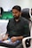 A person is seated in a chair, focused intently on something, with hands positioned on a gaming console or arcade controller. The setup appears to be indoors, with visible screens and lighting in the background, suggesting an electronic gaming environment.