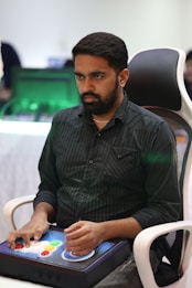 A person is seated in a chair, focused intently on something, with hands positioned on a gaming console or arcade controller. The setup appears to be indoors, with visible screens and lighting in the background, suggesting an electronic gaming environment.